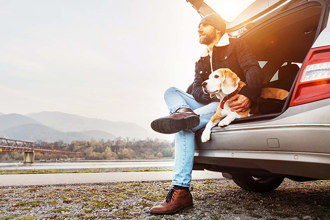 Mężczyzna na postoju w czasie podróży siedzi z psem na krawędzi bagażnika swojego samochodu